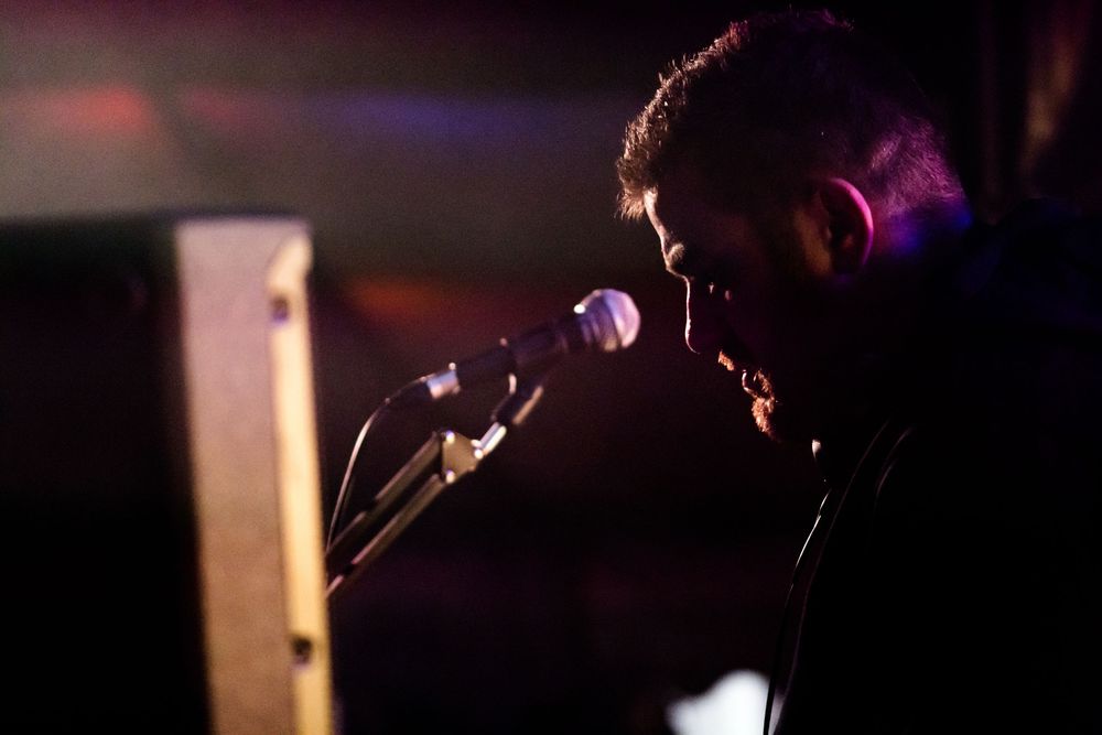 A performer sings passionately into a microphone on a dimly lit stage with dramatic purple lighting in the background.