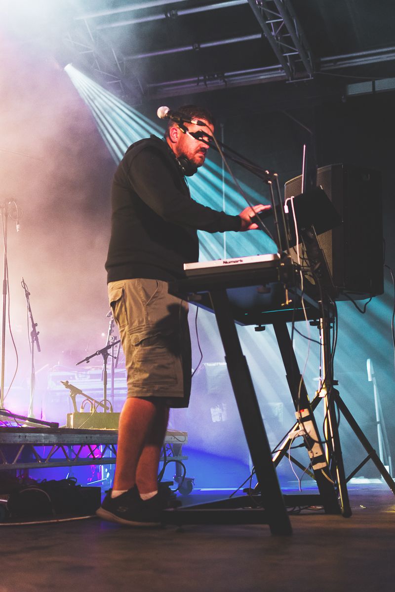 Musician performs on stage with keyboard equipment under dramatic blue and purple stage lighting.
