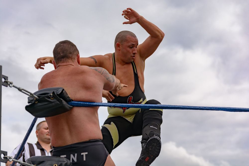 Pro wrestlers engage in a dramatic wrestling match on the top turnbuckle of a wrestling ring under cloudy skies.