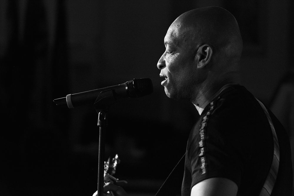 Person performs on stage in black and white with microphone and dramatic lighting in concert setting.