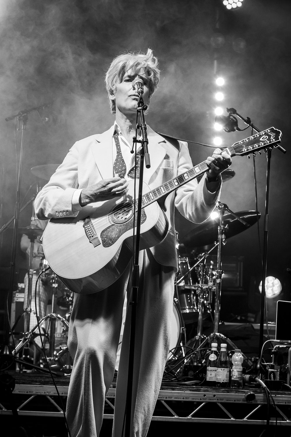 A performer in a white suit plays acoustic guitar on stage under moody lighting and hazy atmosphere at a concert.