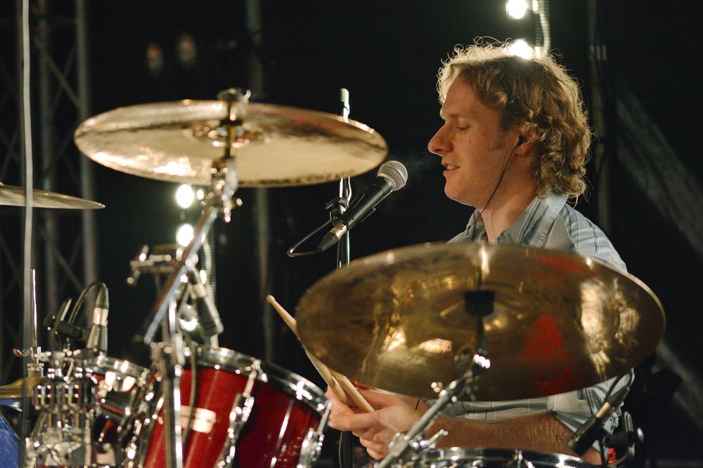 A drummer performs energetically behind a red drum kit with cymbals on a dark stage.