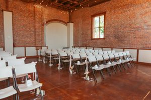 Rows of white chairs arranged in an industrial brick venue with hardwood floors and string lights overhead.