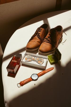 Brown leather shoes, watch, wallet and accessories arranged in dramatic lighting on white surface.