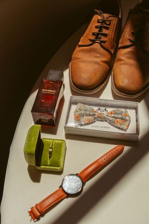 Wedding accessories including brown leather shoes, floral bow tie, ring box, and leather-strapped watch on white surface.