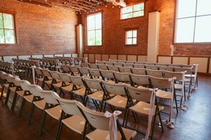 Rows of white folding chairs arranged in a rustic brick-walled venue with large windows.