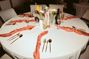 A formal table setting with white tablecloth, coral napkins, gold cutlery, and wine glasses arranged around candlesticks.