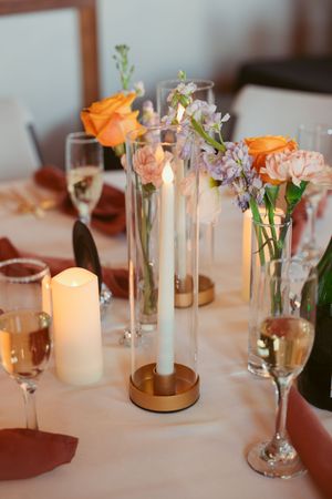 Elegant table setting with tall glass vases holding orange roses and delicate flowers alongside champagne flutes and lit candles.