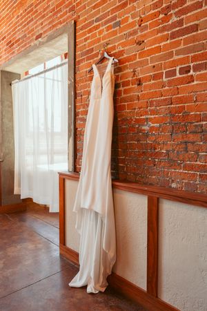 A sleek white wedding dress hangs elegantly against an exposed brick wall next to a sunlit window.