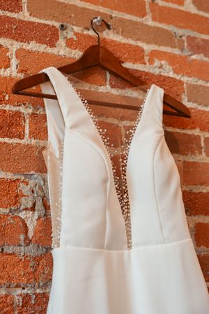 A white wedding dress with plunging neckline and beaded trim hangs on a wooden hanger against exposed brick wall.