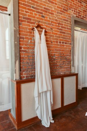 A white wedding dress hangs elegantly against an exposed brick wall with white curtains and wooden wainscoting.