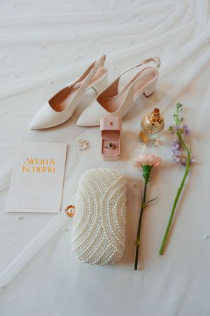 Wedding accessories flatlay with white slingback heels, beaded clutch, perfume bottle, flowers and pink leather belt on fabric.
