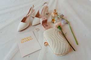 Elegant bridal accessories including blush heels, pearl clutch, gold-toned jewelry, and a wedding invitation on white fabric.