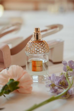 Luxury perfume bottle with gold honeycomb pattern cap displayed with delicate pink and purple flowers on white surface.