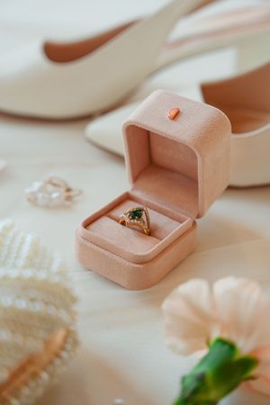 Wedding ring with emerald stone displayed in pink velvet ring box with white heels and pearl accessories in background.