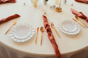 Close-up view of elegantly set dinner table with white plates, rose-colored napkins, and gold flatware on neutral tablecloth.