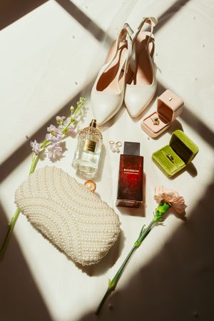 Flatlay of cream bridal shoes, perfume bottles, pearl clutch, and delicate flowers on a white surface with dramatic shadows.