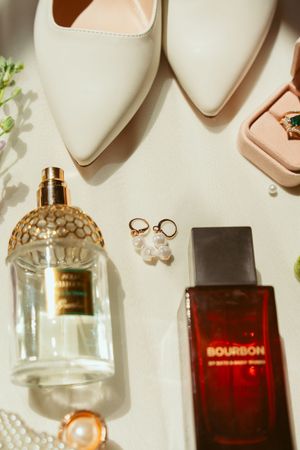 Wedding accessories including pearl earrings, perfume bottles, cream heels and a pink ring box arranged on white background.