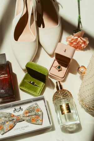 White leather high heel shoes arranged with perfume bottles, velvet jewelry boxes, and delicate pink carnations on a cream surface.
