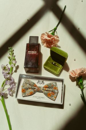 Floral bow tie, perfume bottles, and delicate purple flowers arranged on white surface with diagonal shadow patterns.