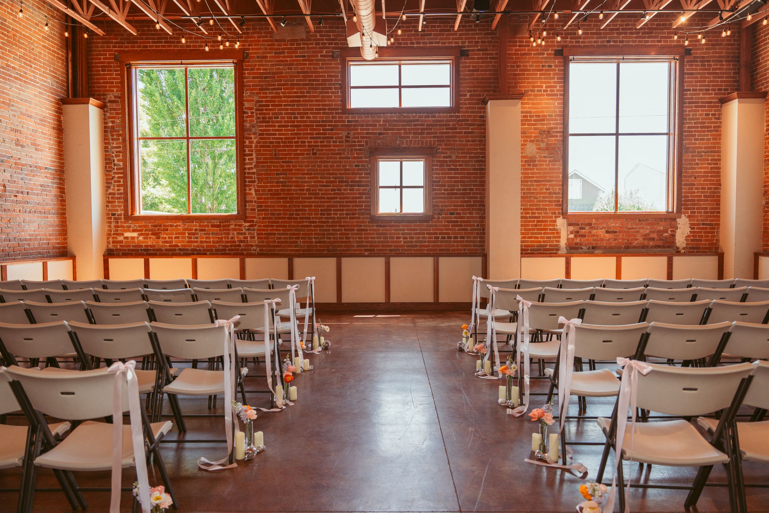 Modern industrial style wedding ceremony venue with exposed brick walls, gray chairs in rows, and natural light through windows.