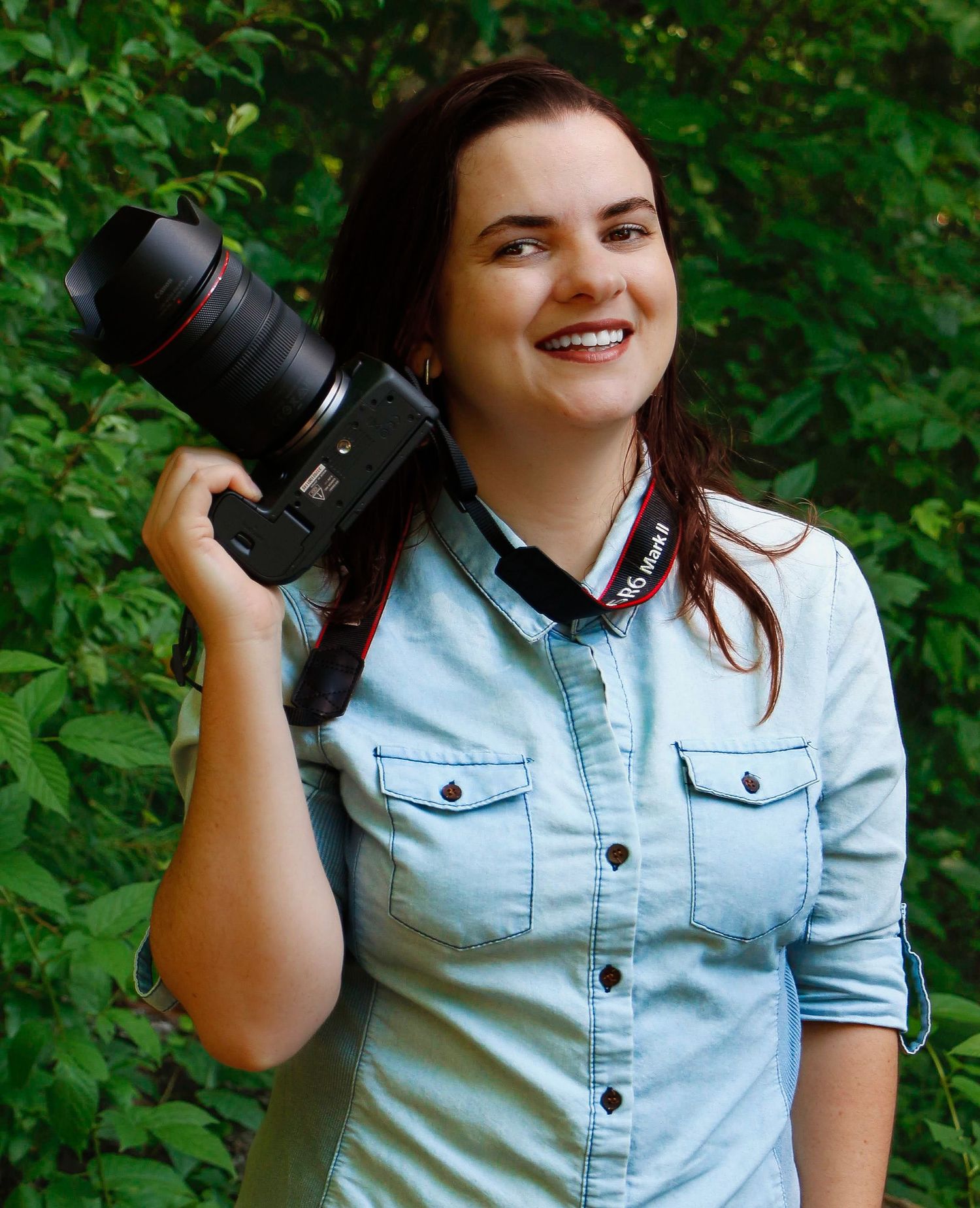 Photographer displays professional camera equipment while wearing denim shirt against green foliage.