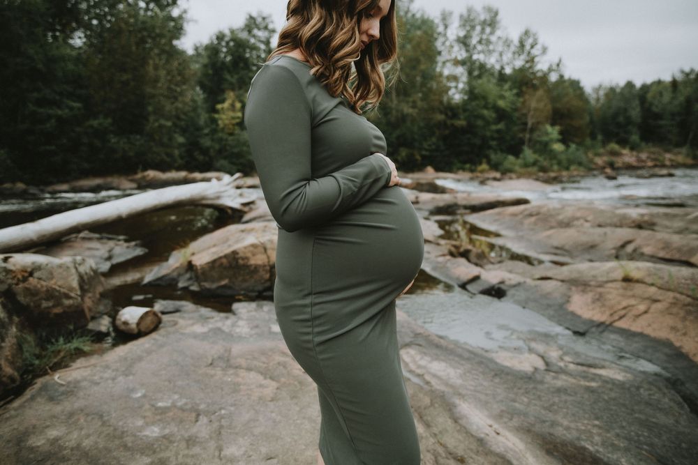 Side view of pregnant woman in green dress posing on rocky terrain near water.