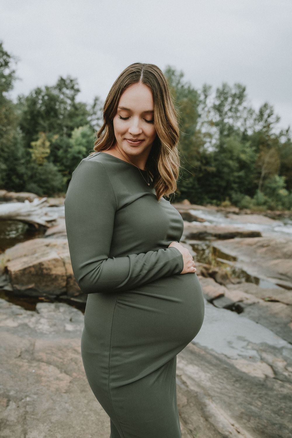 Maternity photo of woman in olive green dress standing on rocky outcrop by water.