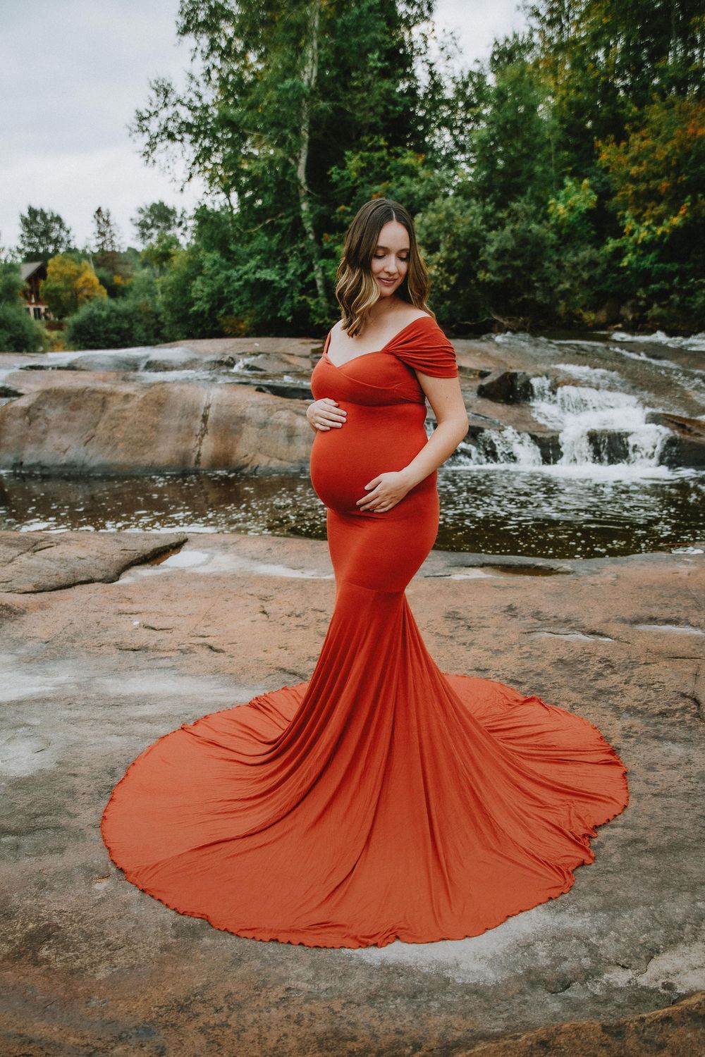 Maternity photo featuring woman in flowing red dress standing near waterfall on rocky terrain.