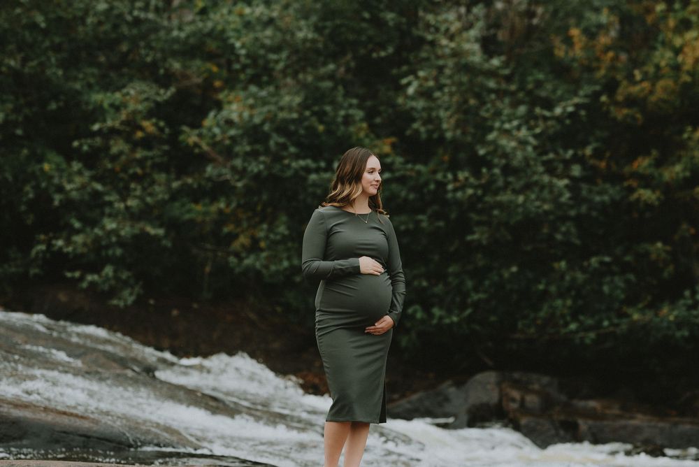 An expectant mother in an olive green dress posing on rocky terrain.