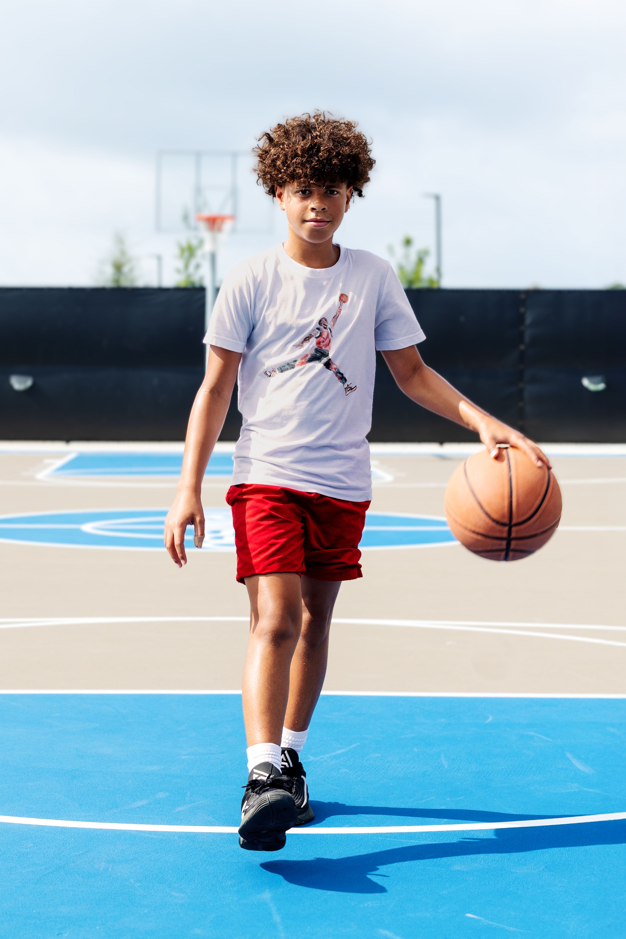 Young basketball player dribbling on an outdoor court wearing a white t-shirt and red shorts.