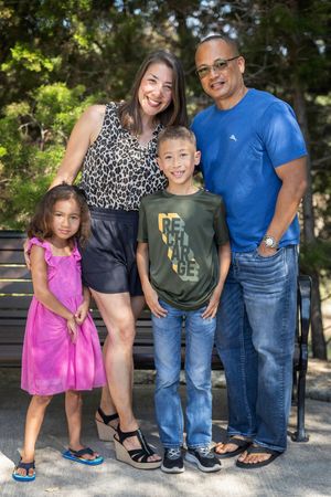 Family of four smiling together while posing outdoors, wearing casual clothing on a sunny day.