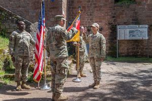 Group of military service members stand in formation with American flags during an outdoor ceremony near brick wall.