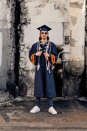 Graduation portraits taken in urban setting showing person in navy blue cap and gown with orange stole against industrial wall.