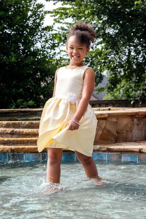 A child in a yellow and white dress splashes their feet in a poolside water feature on a sunny summer day.