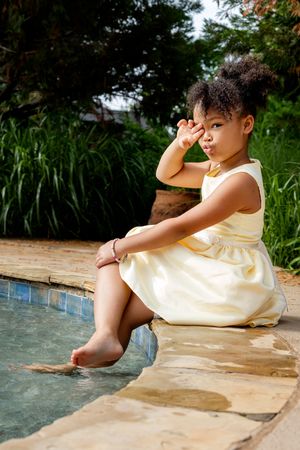 Child in cream-colored dress sits at edge of swimming pool on sunny day surrounded by lush garden landscaping.