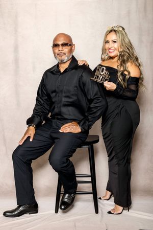 A couple poses together in elegant black attire against a light background during a formal photoshoot.
