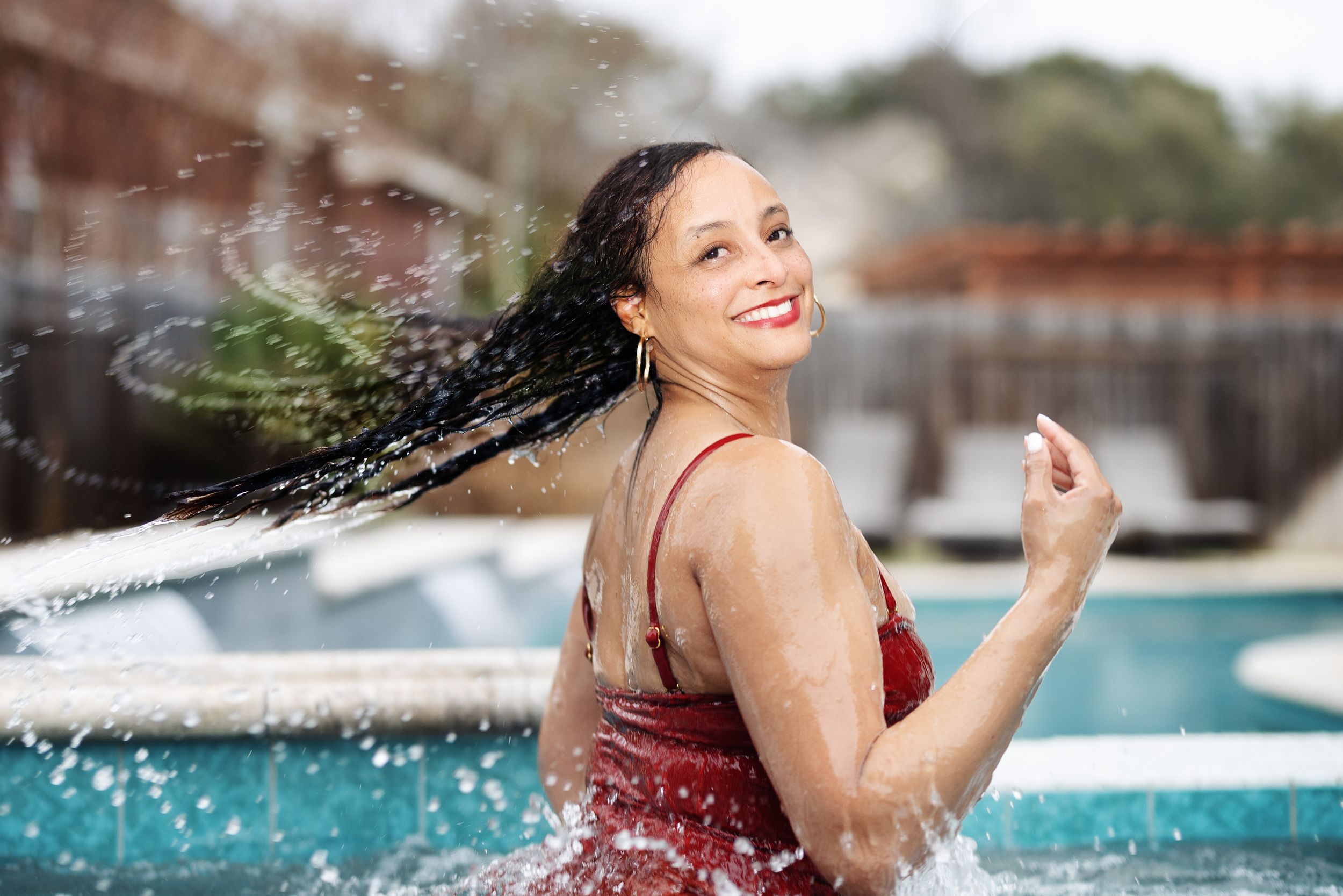 Woman in red swimsuit flipping wet hair by a pool, smiling joyfully with water splashing around her.
