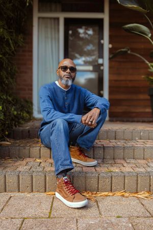 A person in a blue sweater and jeans sits casually on brick steps outside a house wearing brown leather high-top sneakers.
