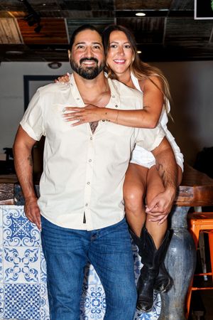 A couple in coordinating white shirts and blue jeans embracing and smiling in front of a wall with blue tile accents.