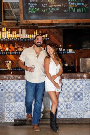Couple poses at rustic bar with blue and white tiled backsplash and chalkboard menu overhead.