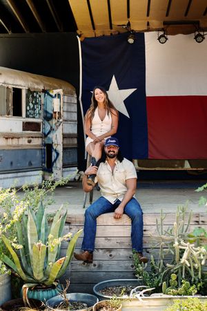 A couple sits on rustic wooden steps in front of a Texas state flag with potted succulents and agave plants in the foreground.