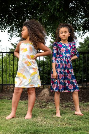 Two children in summer dresses dancing and playing together on a grassy lawn near a black fence.
