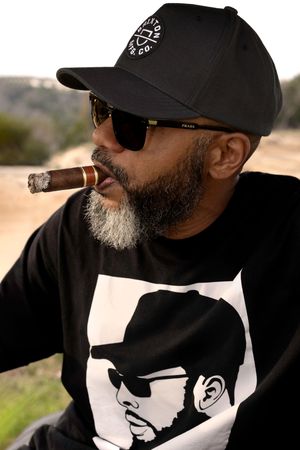 Person in black baseball cap and shirt smoking cigar outdoors, wearing sunglasses with graphic design on their clothing.