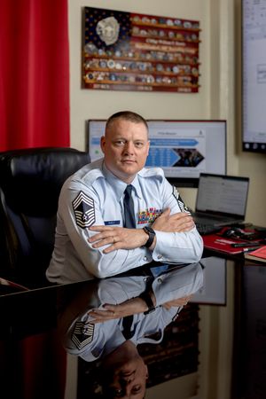 A military service member in dress uniform sits at a desk with a baseball memorabilia display on the wall behind them.