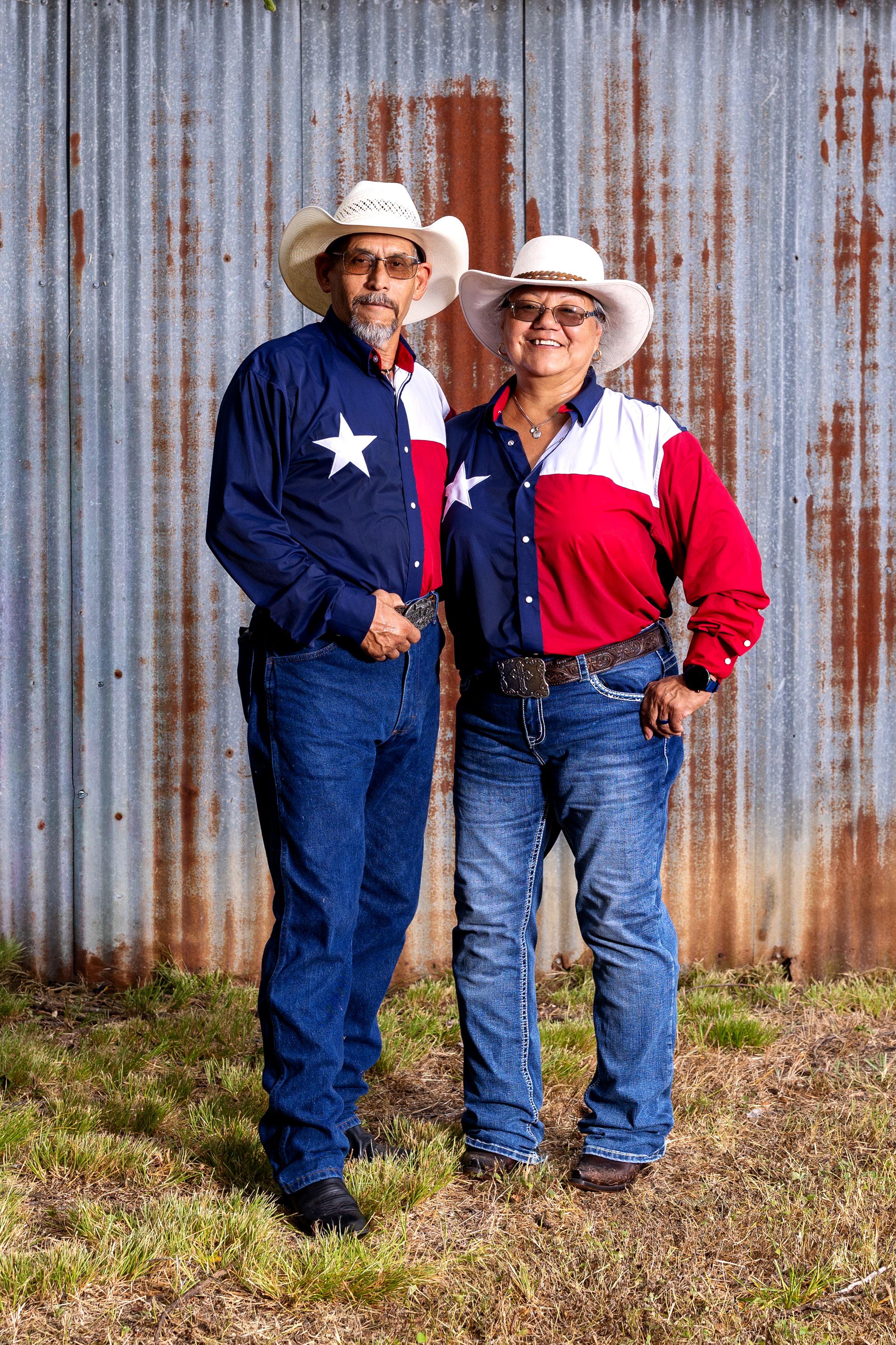 Couple wearing matching Texas flag shirts and cowboy hats posing in front of a rustic corrugated metal wall.