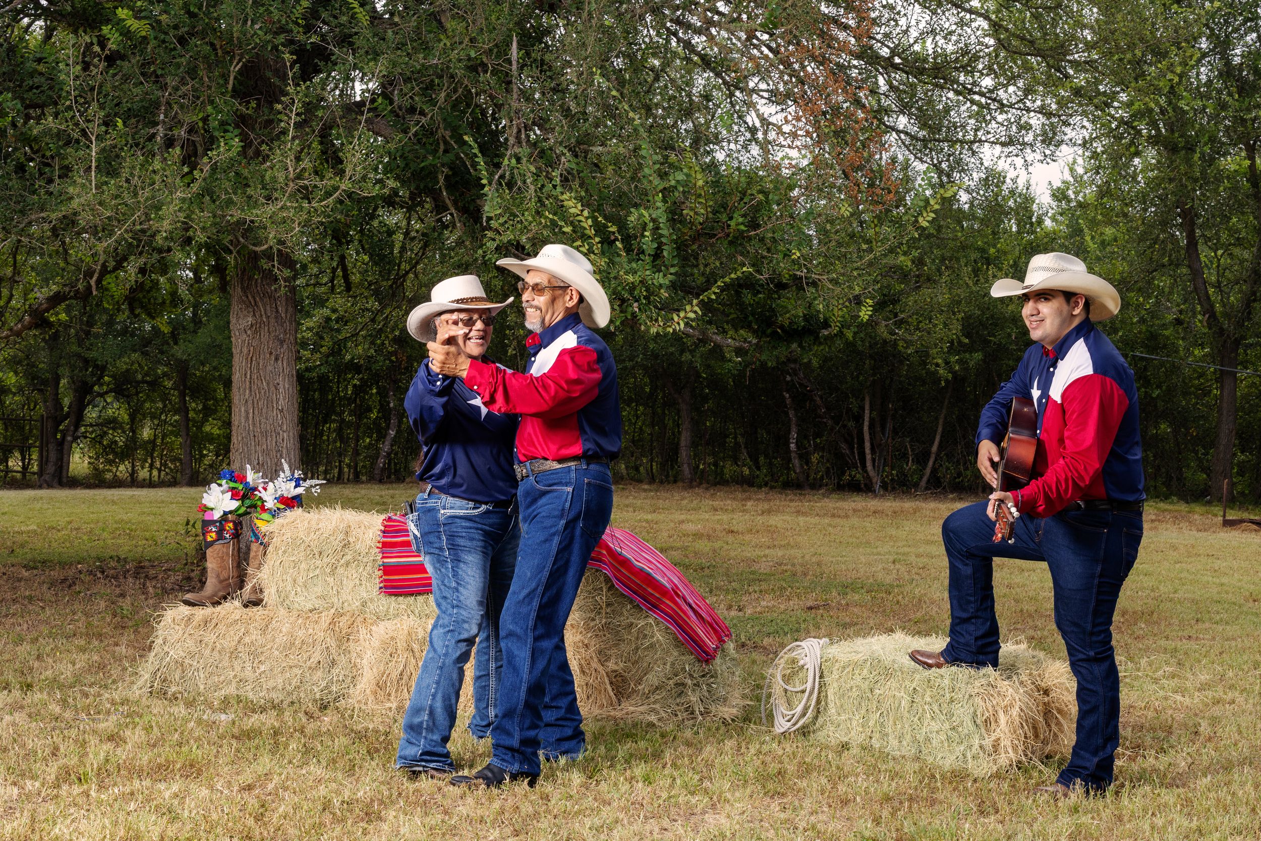 Three people in cowboy hats and red and blue outfits pose near hay bales outdoors, one playing guitar.