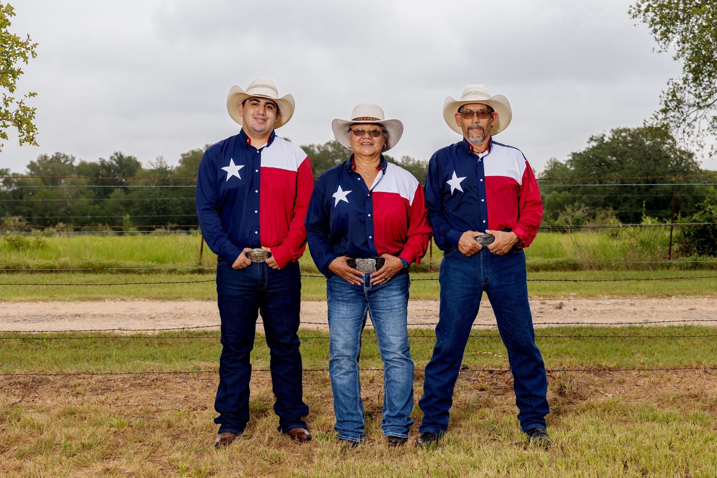 Three people in matching Texas flag cowboy outfits posing outdoors on a dirt path with green fields behind them.