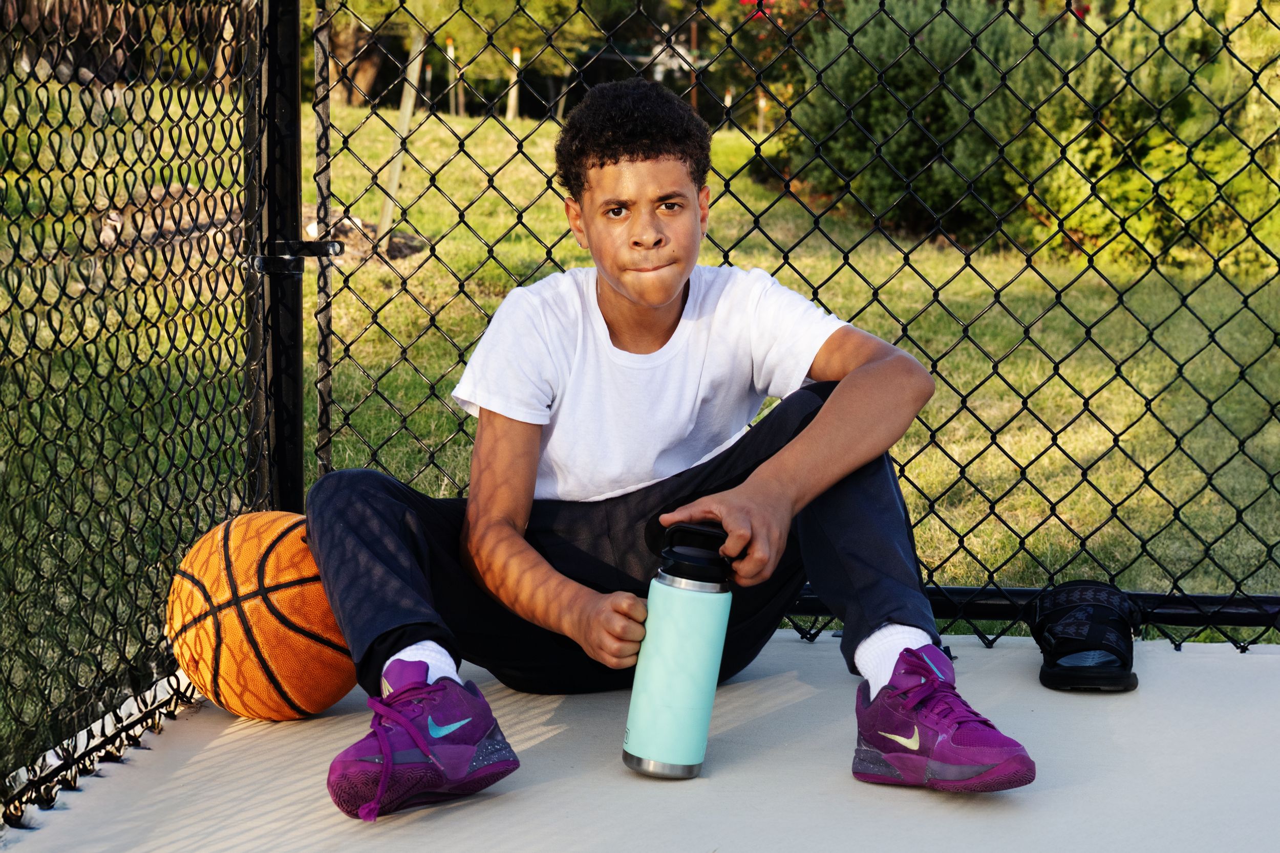 Young boy sitting on outdoor basketball court holding water bottle next to orange basketball, wearing purple sneakers.