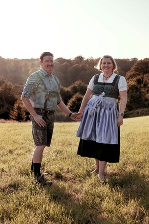A couple in traditional Bavarian dirndl and lederhosen dance together in a sunlit meadow during golden hour.